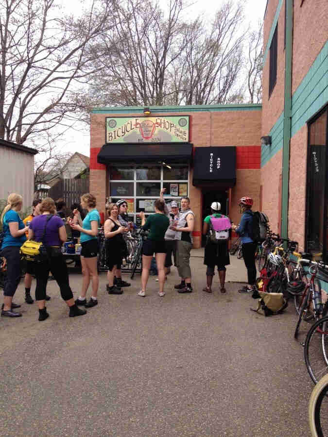 A group of cyclist, gathered in front of a bike shop, with their bikes leaning against the building walls around them