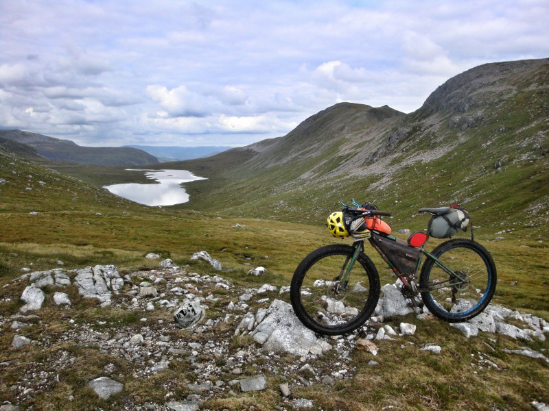 Left side view of a green Surly bike with gear, on a rock patch in a mountain valley, with a lake in the background