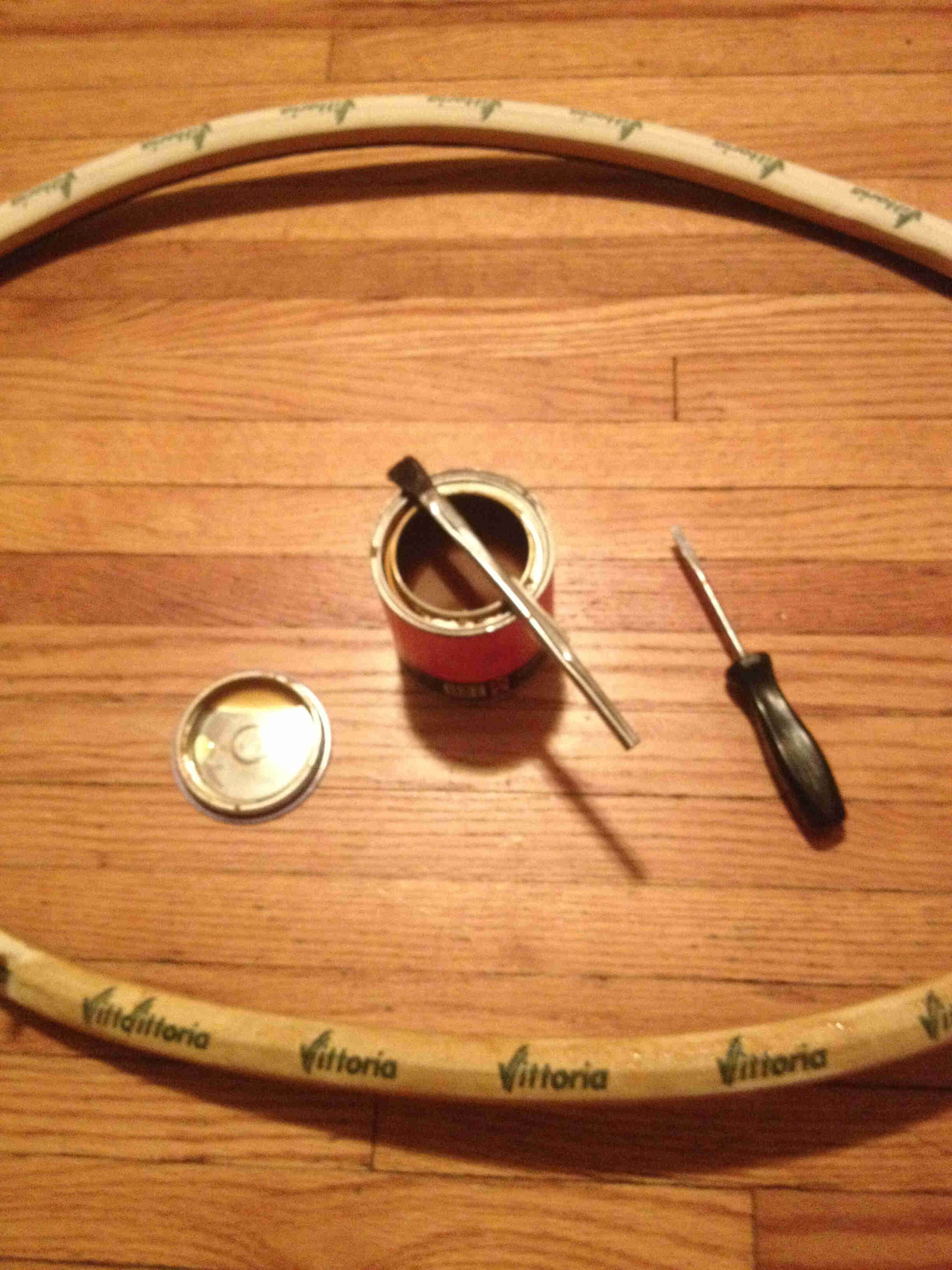 Downward view of a Vittoria bike tire, with a can and brush in the middle,  laying on a wood floor