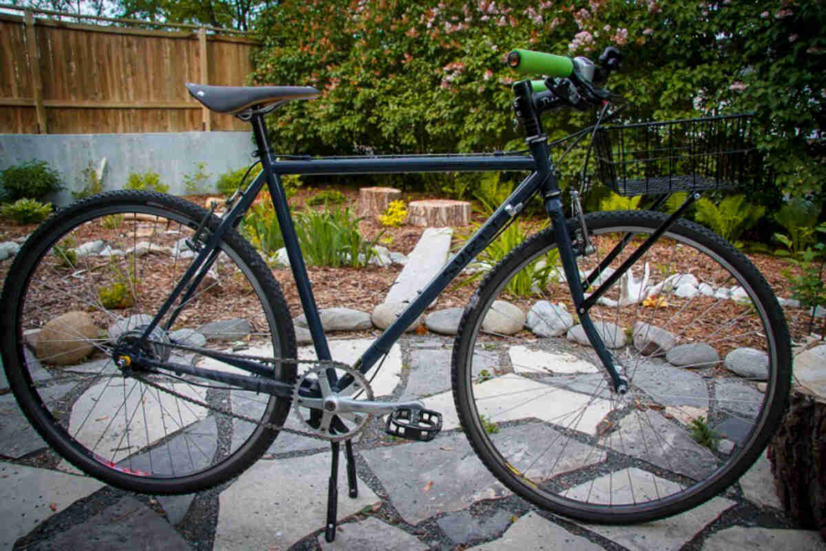 Right side view of a black Surly Cross Check bike, on a stone patio, with landscaping, bushes and a wood fence behind