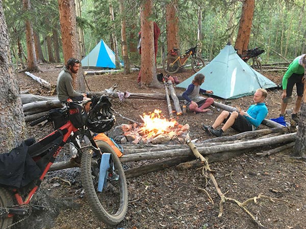 People at camp in woods, loaded bikes parked, tents set-up and campfire burning