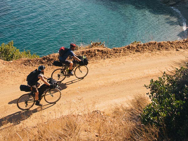 View looking down, two cyclists wearing helmets and backpacks riding loaded drop bar bikes on gravel road next to ocean