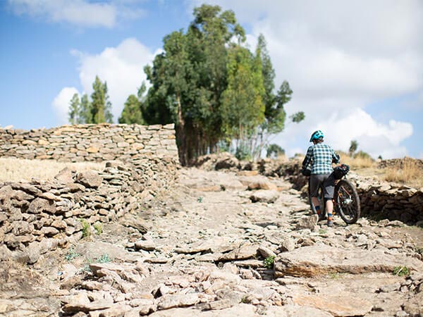 Person pushing loaded bike up rocky trail next to stacked stone walls