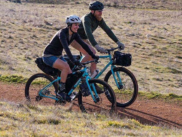Two cyclists wearing helmets riding loaded Bridge Club bikes on gravel road through grasslands