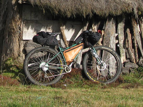 Green Surly Krampus loaded with bags leaning against wooden shelter with sod roof on sunny day