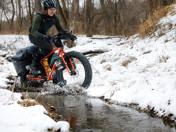 Person riding loaded orange Pugsley off-road through small stream in woods, snow on ground, tires splash through water