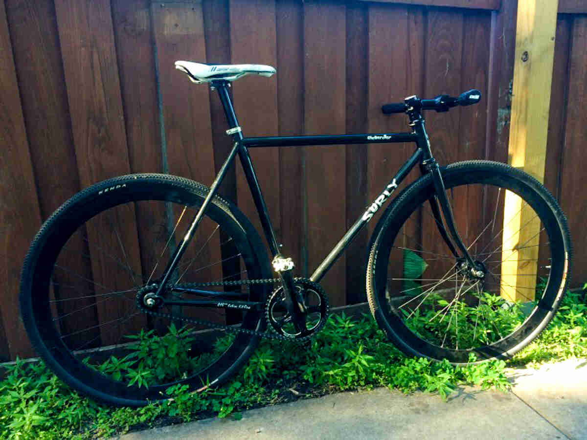 Right side view of a black Surly Steamroller bike, leaning against a wood fence wall