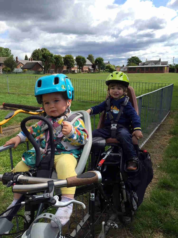 Front view of 2 children in bike seats mounted on a black Surly Big Dummy bike, parked against a fence in a grass field