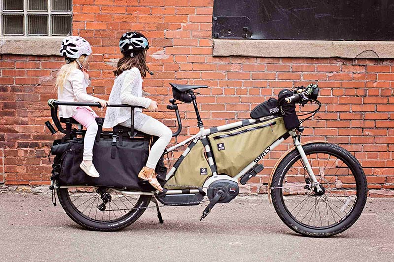 Right side view of a white Surly Big Easy bike with rear carrier holding two small children, next to a brick building