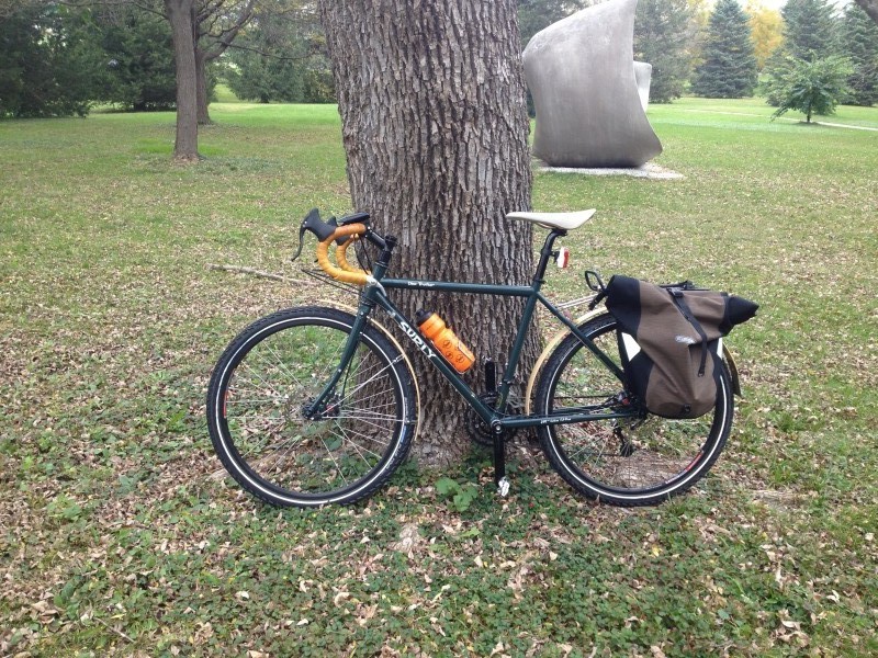 Left side view of a Surly Disc Trucker bike, leaning on a tree in a grass field, with a sculpture in the background