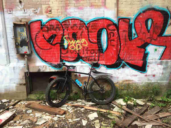 Left side view of a black bike, parked on building debris, against a brick wall with graffiti
