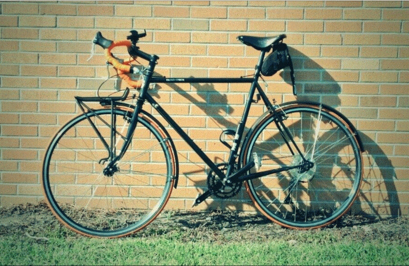 Left side view of a black Surly Cross Check bike, parked on grass and leaning against a brick wall