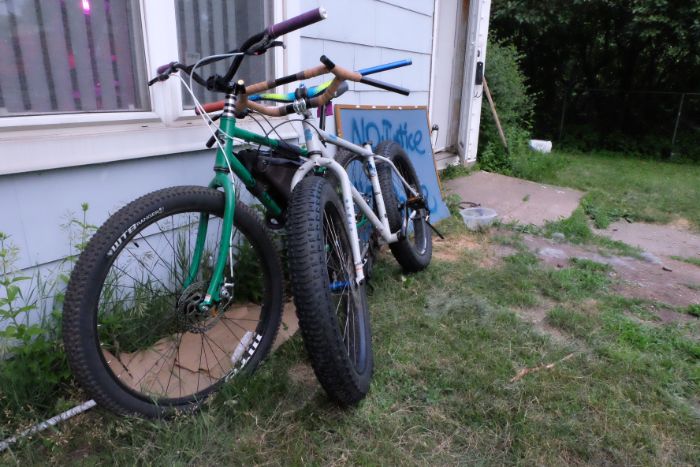 One green fat bike and one white fat bike with no seat lean on each other against a light blue home 