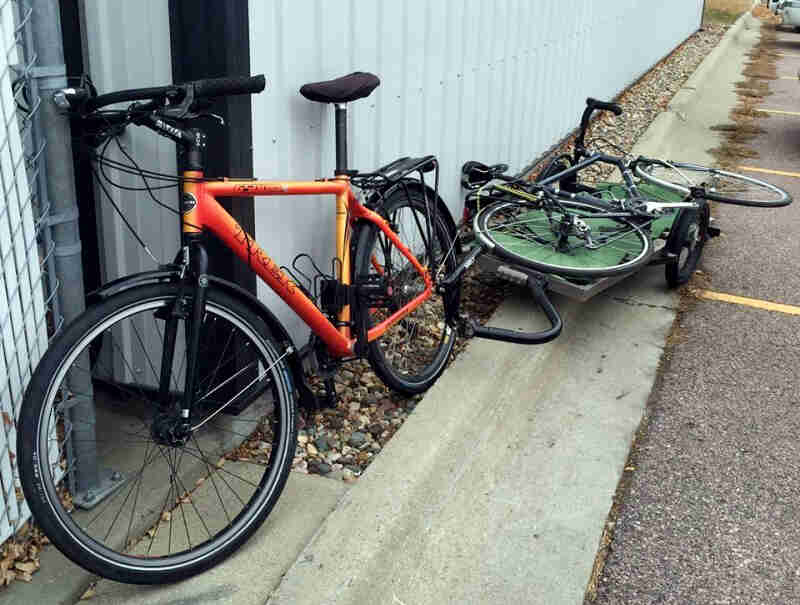 Right front view of an orange bike with a trailer hauling a bike hitched behind, parked against a steel building