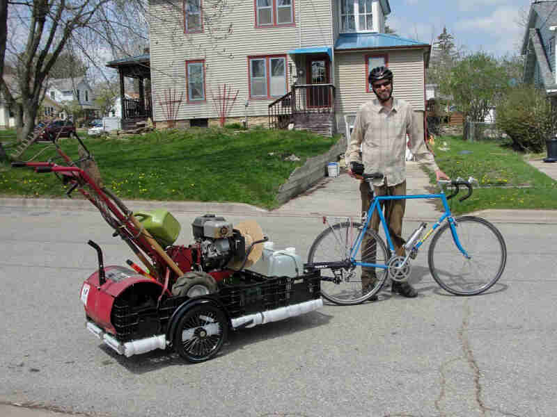 Cyclist standing on a street with a Surly bike, blue, with a trailer hauling a rototiller attached to the back