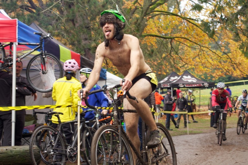 Front, left side view of a cyclist, wearing shorts and sticking their tongue out, riding a Surly bike on a muddy trail