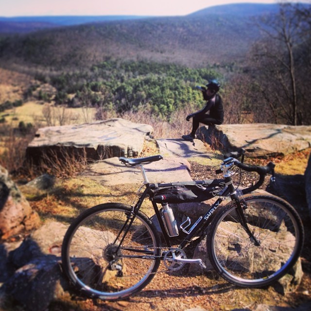 Right side view of a black Surly Cross Check bike, on a rock hilltop with a cyclist sitting behind, in the mountains