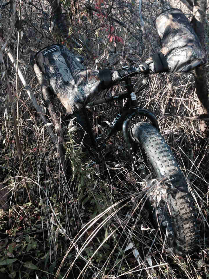 Front view of a black Surly Pugsley fat bike with camo hand warmers, parked in heavy brush in the thick, bare woods