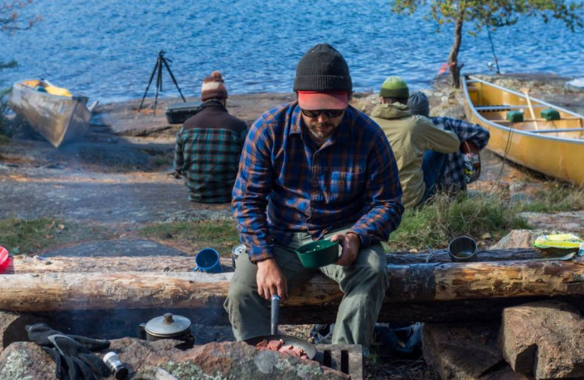 Front view of a person sitting on a log at a campsite, cooking, with people and a lake in the background