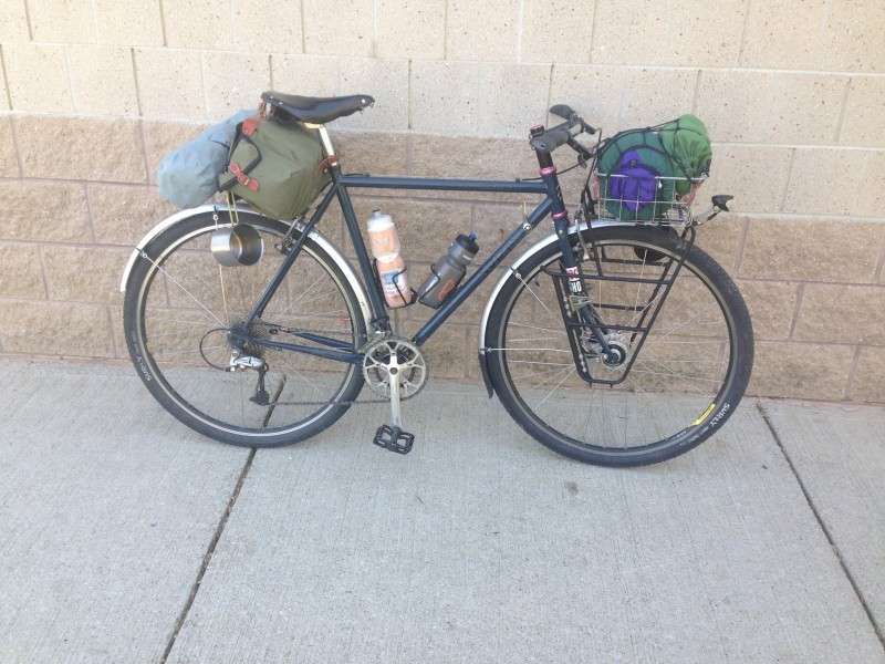 Right side view of a teal Surly Cross Check bike with gear packs, parked on a sidewalk, leaning on a stone block wall