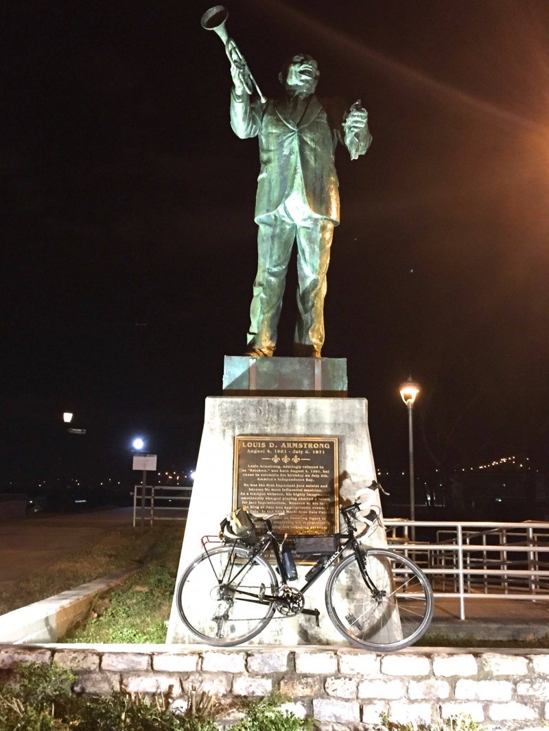 Right side view of a black Surly Cross Check bike, leaning on a stone base of a bronze Louis Armstrong statue at night