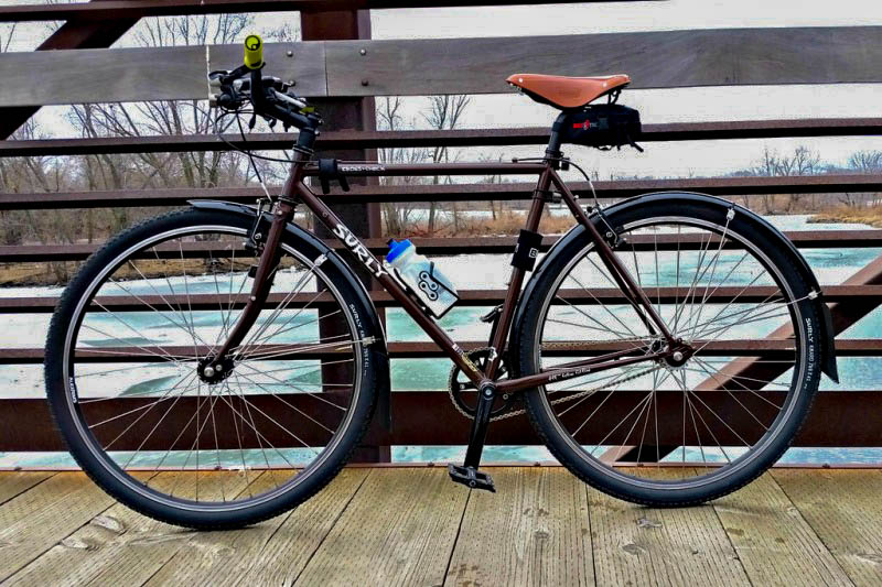 Left side view of a Surly Cross Check bike, brown, parked on a bridge over a river