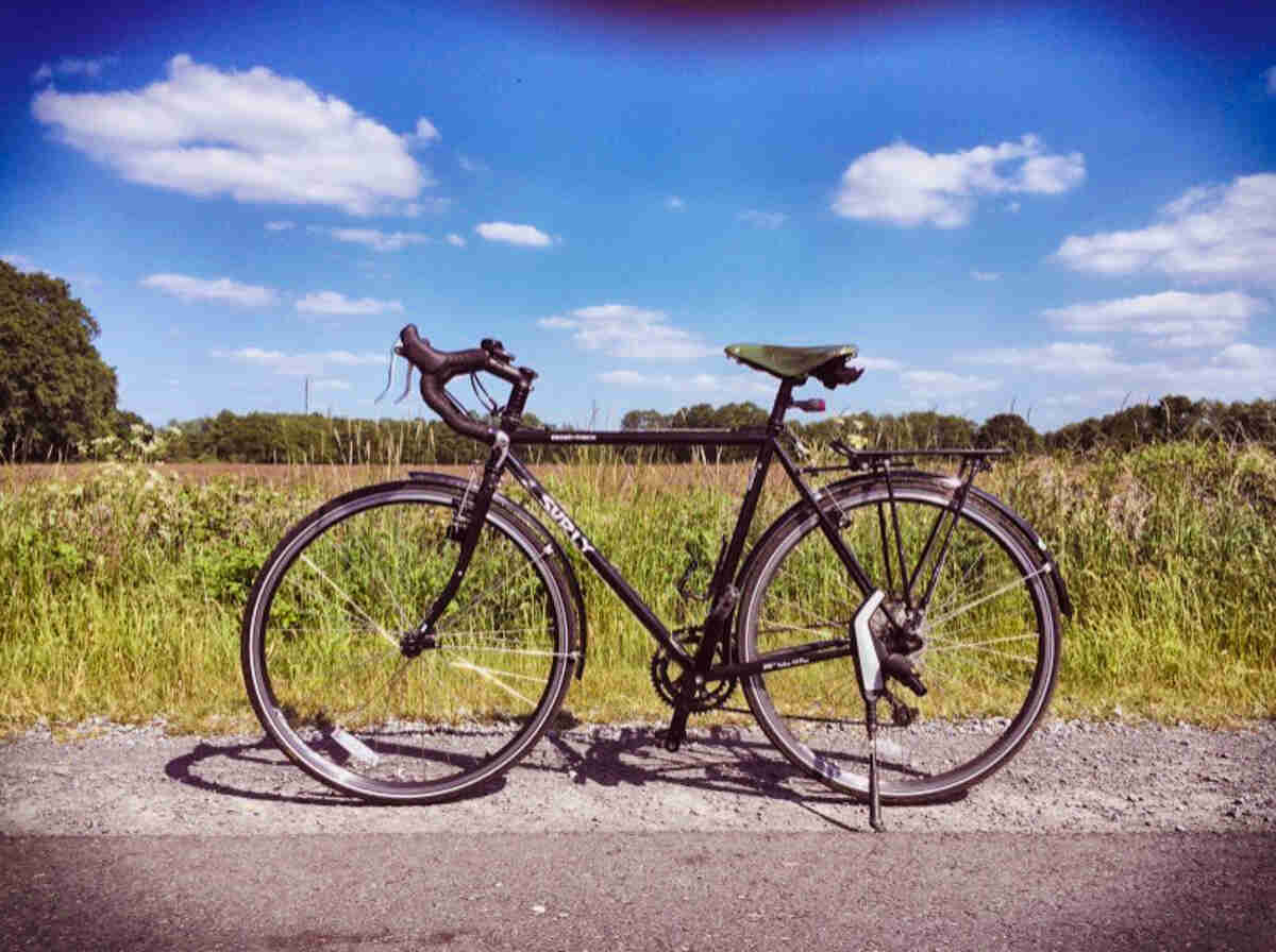 Left side view of a black Surly Cross Check bike, parked on a roadside, with a tall grass field in the background