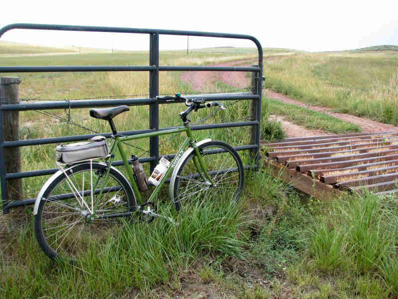Right side view of a green Surly bike with a seat pack, parked in grass against a gate, next to a dirt road in a field