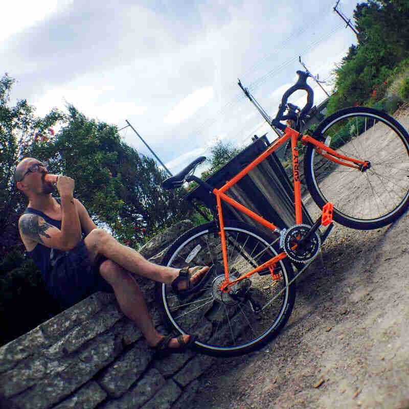 Tilted right side view of an orange Surly bike, and a cyclist sitting on a stone wall behind the back end, in a dirt lot