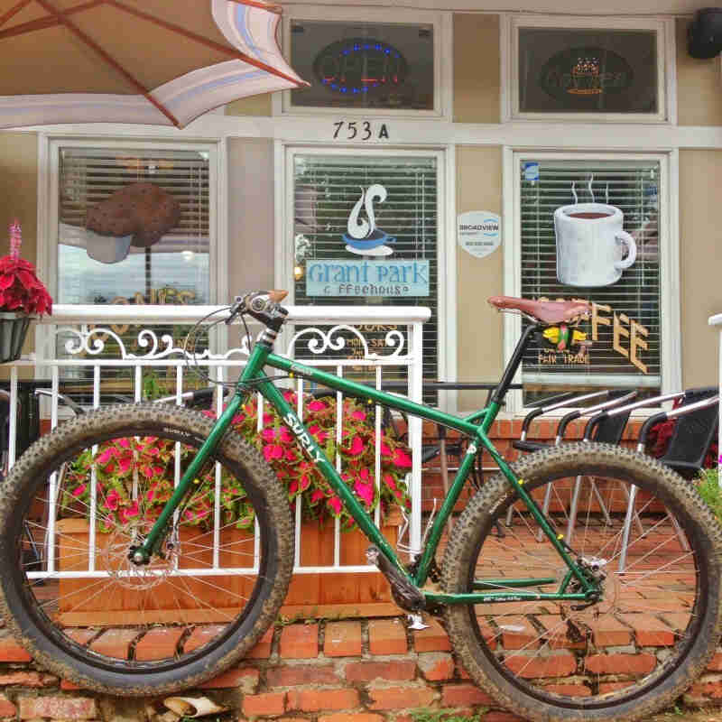 Left side view of a Surly Krampus bike, green, on a red brick sidewalk in front of a coffee shop