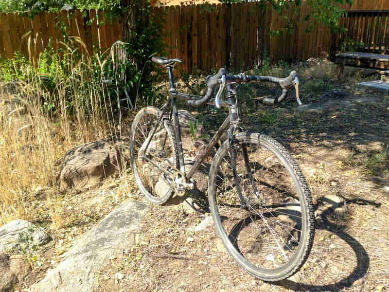 Front, right side view of a black Surly Cross Check bike, parked on dirt and rocks, with weeds and a wood fence behind