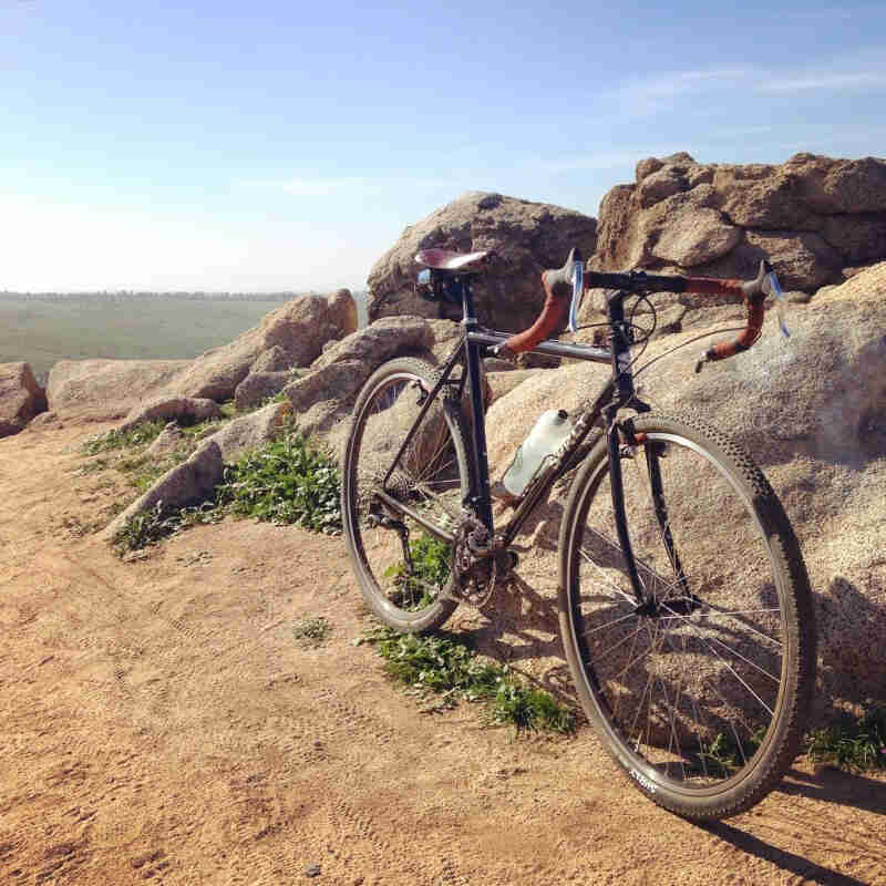 Front, right side view of a black Surly Cross Check bike, parked on dirt, next to a boulder mound