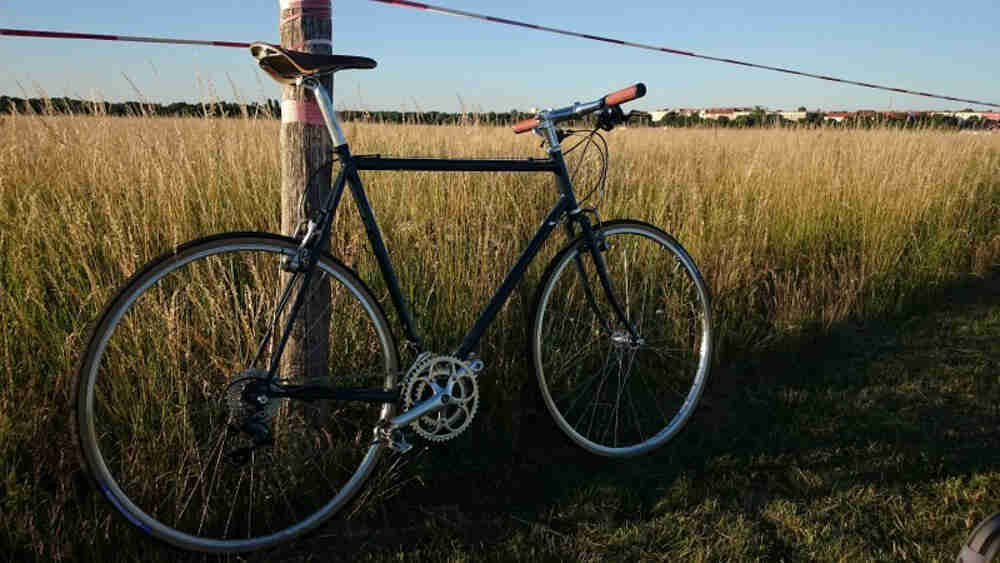 Right side view of a black Surly Cross Check bike, parked in grass against a wood post, with a tall grassy field behind