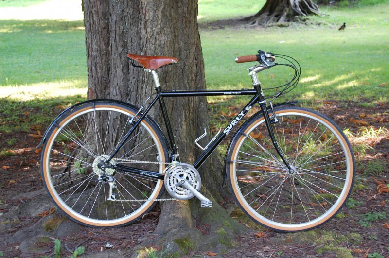 Right side view of a black Surly Cross Check bike, parked in front of the base of a tree, with grass field behind