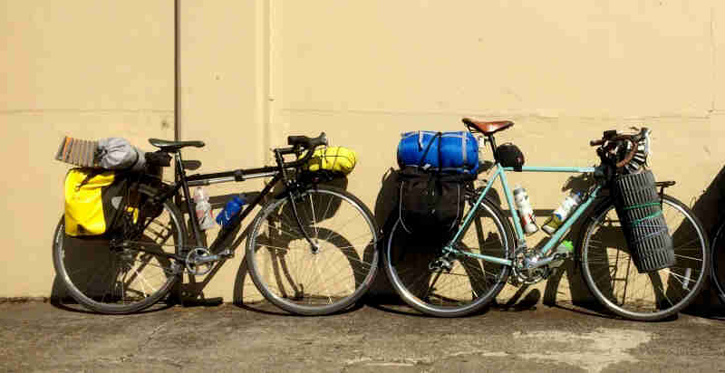 Right side view of 2 Surly Cross Check bikes loaded with gear, parked single file against a yellow cement wall