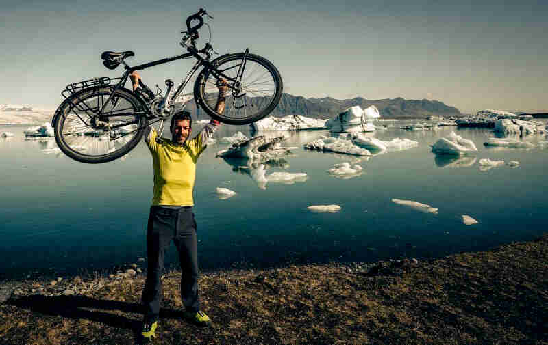 A cyclist holds up a black Surly bike, on the shore of turquoise waters, with icebergs floating around