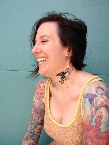Right side view of a smiling person, from the waist up, with a tattoos on their neck and arms