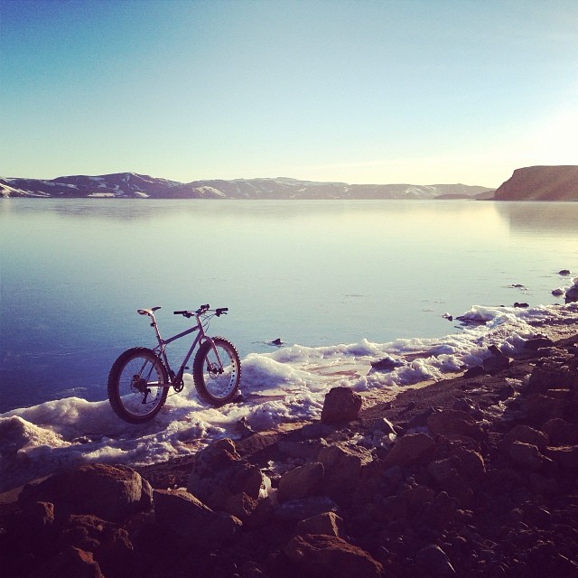 Right side view of a Surly fat bike, on an icy bank of a lake, with snowy mountains around it