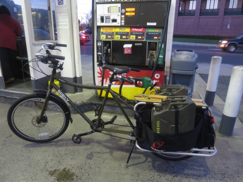 Left side view of an olive color Surly Big Dummy bike with diesel cans on back, parked alongside a fuel, pump station