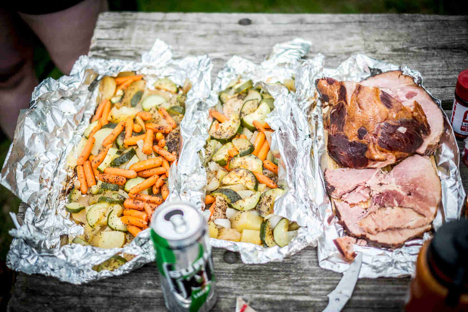 Downward view of 2 sheets of foil with cooked vegetables in them, and a beer can in the forefront, on a table