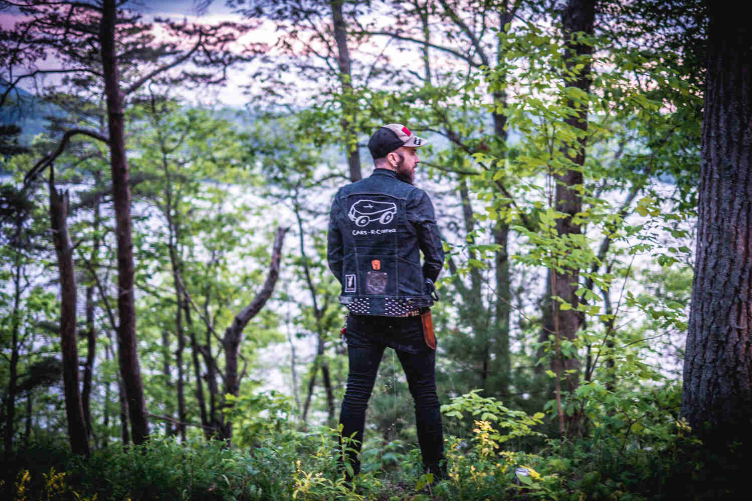 Rear view of a person standing while facing trees, wearing a jean jacket with, Cars-R-Coffins, spelled on the back