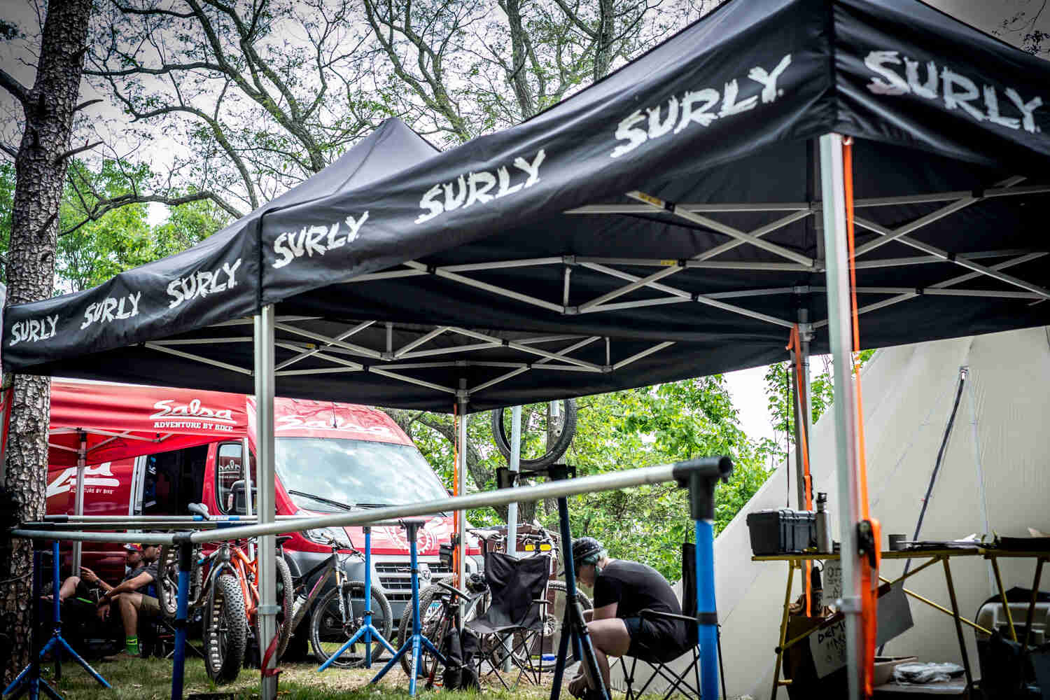 Cyclists and bikes, under a Surly bikes canopy in front of a red van, in the woods