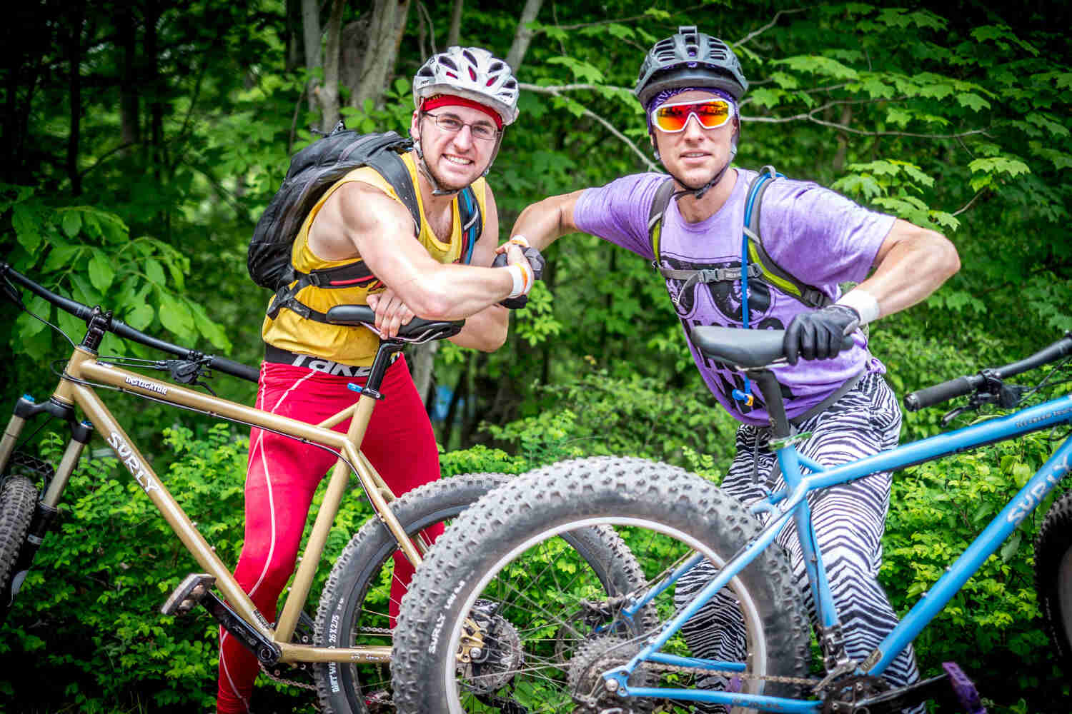 Left side view of a tan Surly Instigator bike with 2 cyclist posing behind, shaking hands, and woods in the background