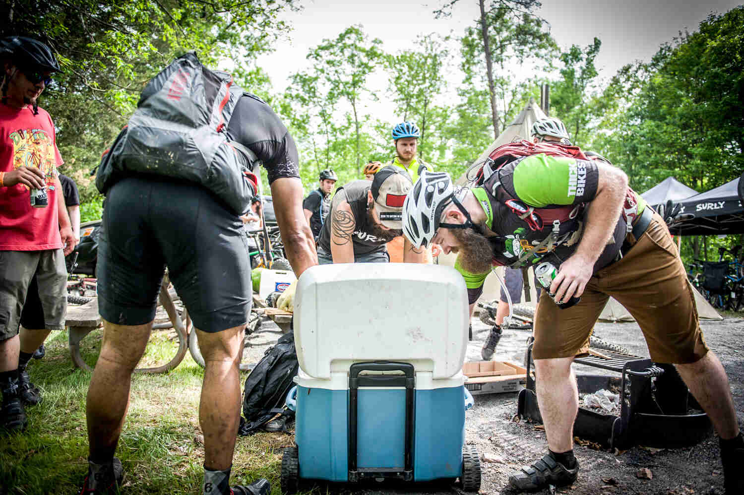 A group of cyclists, reaching into a cooler, at a campsite in the woods