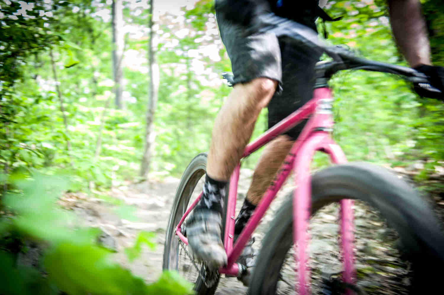 Blurred, front right side view of a cyclist, riding a pink Surly bike on a dirt trail in the woods