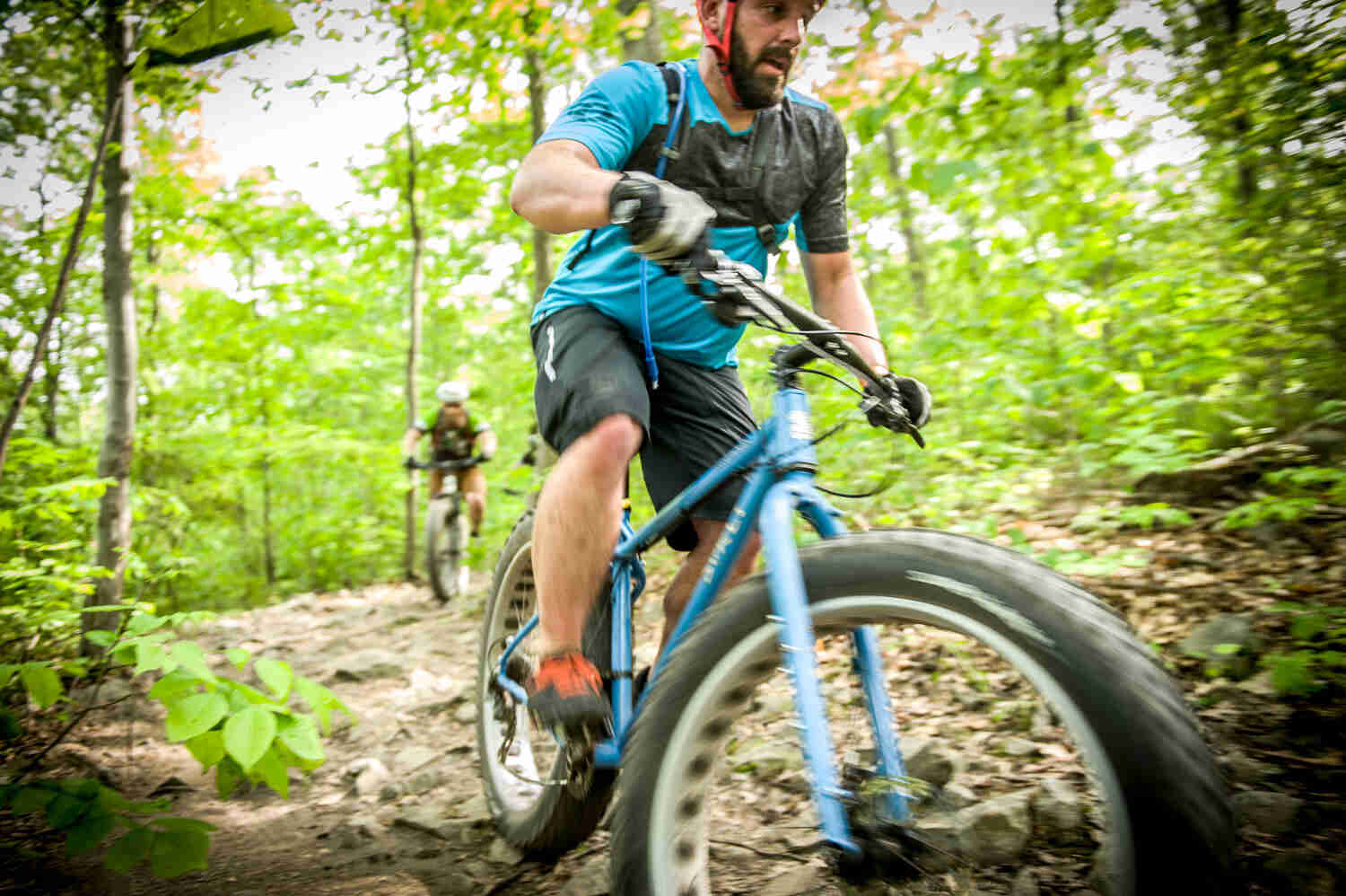 Front view of a cyclist, riding a blue Surly Ice Cream Truck fat bike, on a rocky trail in the woods