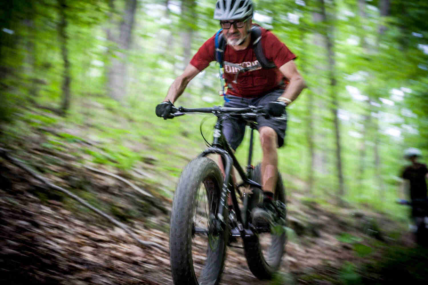 Blurry, front left side view of a cyclist riding a black Surly fat bike, on a trail in the green woods