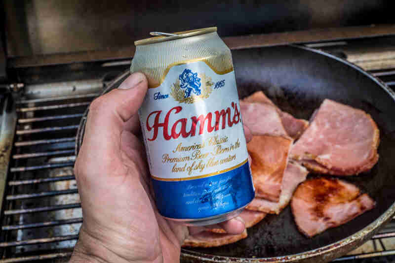 A hand holding a can of Hamm's beer over a frying pan of fried ham, above a grill grate