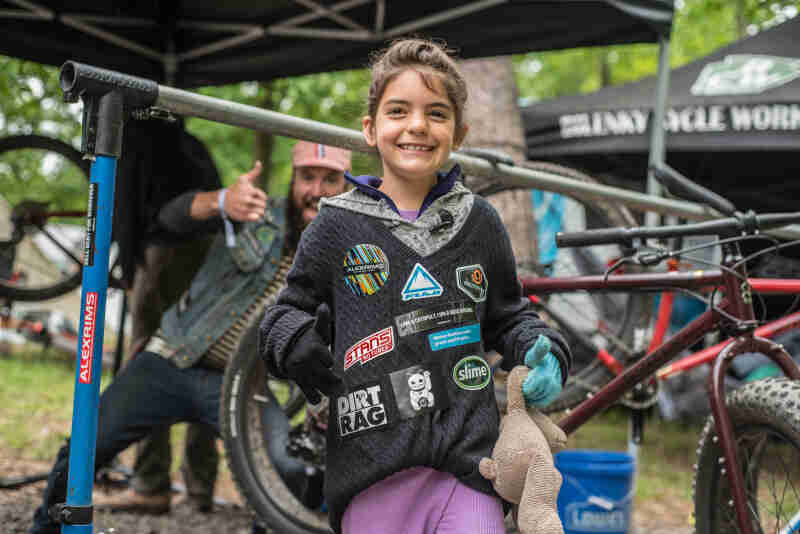 Front view of a child smiling, with a bike and person giving a thumbs up in the background