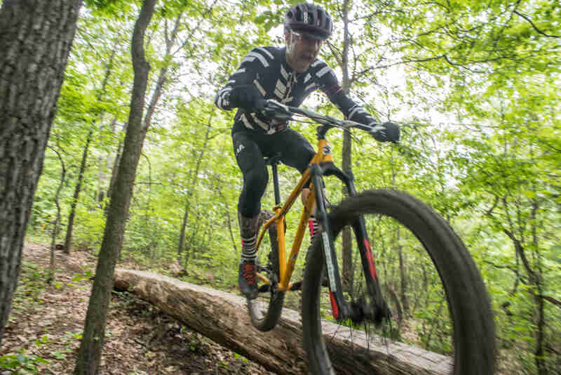 Front right side view of a cyclist riding down a log bridge on a hill in the woods, on a yellow Surly bike